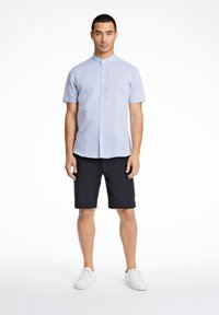 Camicia azzurra a maniche corte con colletto a band, abbottonata, abbinata a pantaloni corti blu scuro. Modello che indossa sneaker bianche, in piedi su uno sfondo neutro.