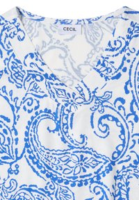 Vêtement blanc avec motifs floraux paisley bleus et col en V, étiqueté "CECIL" sur une étiquette cousue.