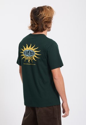 Homme aux cheveux châtain clair porte un t-shirt Volcom vert foncé avec un soleil jaune et un logo au dos, associé à un pantalon marron foncé.
