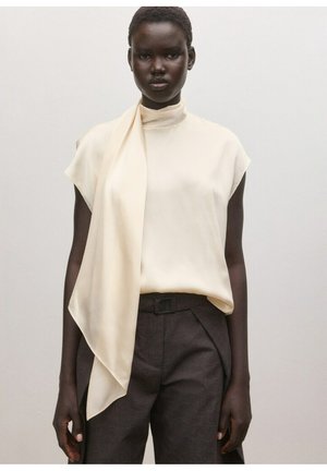 Kobieta o ciemnej karnacji, ubrana w kremową, bezrękawnikową bluzkę z asymetrycznym szalikiem oraz ciemnoszare spodnie z wysokim stanem.