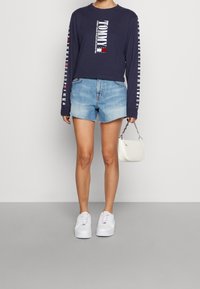 Námořnická modrá košile s dlouhým rukávem a bílým a červeným logem Tommy, spárovaná se světle modrými denimovými šortkami a bílými teniskami. Drží malou bílou tašku.