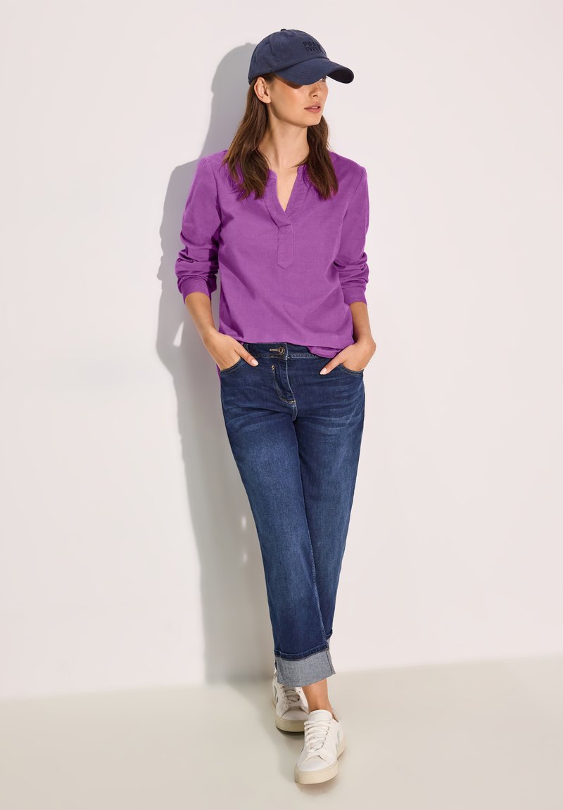Lila långärmad skjorta med krage, kombinerad med mörkblå jeans och vita sneakers. En marinblå keps fulländar outfiten.