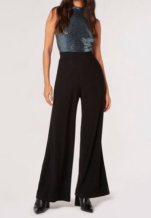 Femme portant un pantalon noir taille haute à jambes larges, un haut bleu brillant sans manches et des bottines noires à talons, debout devant un fond uni.