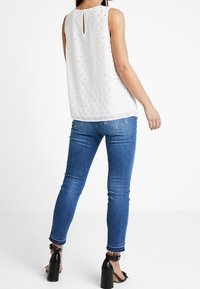 Vit ärmlös topp med strukturerat prickmönster, detalj med nyckelhål bak, tillsammans med smala blå jeans och svarta klacksandaler.