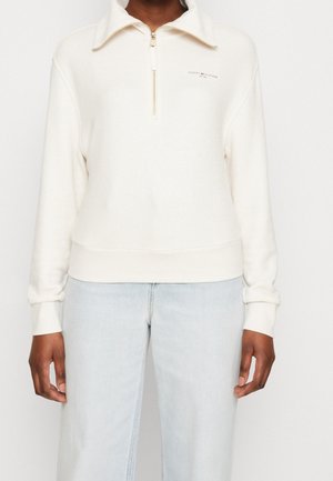 Kvinde iført en hvid kvart-zip sweatshirt med et lille logo på brystet og lyseblå jeans, stående foran en ensfarvet baggrund.