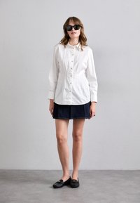 Witte button-up shirt met een op maat gemaakte pasvorm, denim minirok en zwarte instappers. Gecombineerd met een donkere zonnebril en een neutrale achtergrond.