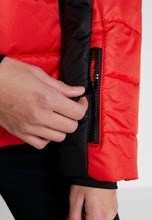 Hand stelt zwarte ritssluiting op rode gewatteerde jas mouw met zwarte manchet eronder af tegen een neutrale achtergrond.