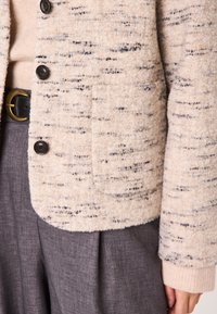 Veste beige texturée en tissu avec des éclats sombres, dotée d'une coupe carrée, de cinq boutons noirs et d'une poche à rabat sur le devant.