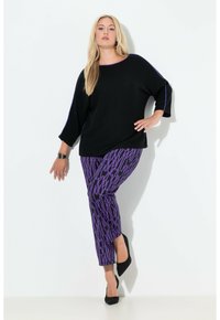 Czarna, oversize bluzka z fioletowymi akcentami, zestawiona z fioletowymi spodniami w wzory z czarnym nadrukiem. Modelka ma na sobie czarne szpilki.