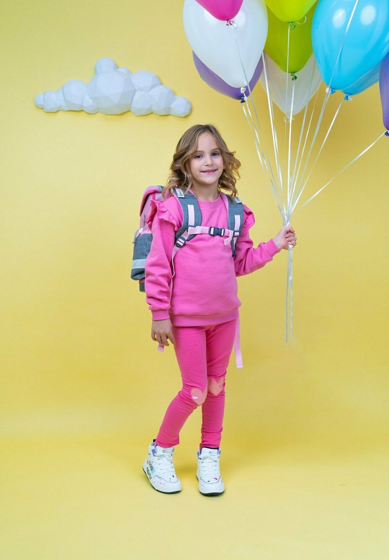 Rosa Sweatshirt und Leggings, grauer Rucksack, hält bunten Ballons. Hintergrund ist gelb mit einer weißen Wolke. Sneaker mit Akzenten.
