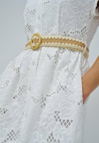 Vestido blanco de encaje floral con botones en la parte delantera, un cinturón beige texturizado con una hebilla redonda y una mano metida en el bolsillo.