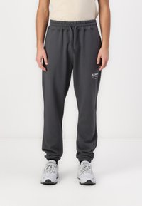 Kohlgraue Sweatpants mit elastischem Bund und Kordelzug, gerippten Bündchen und einem kleinen weißen Logo an der linken Oberschenkelseite.
