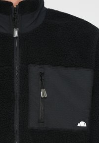 Juodas vilnonis švarkas su aukšta apykakle, turintis krūtinės kišenę su užtrauktuku ir tekstūruotą audinį. Matomas baltas logotipas ant kišenės srities.