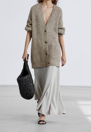 Femme marchant, portant un cardigan en maille beige oversize boutonné, une jupe légère fluide, des sandales noires, et portant un grand sac tissé noir.