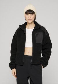 Černá fleecová bunda se zapínáním na zip vpředu a texturovaným povrchem. Má černé nylonové panely a zipovou kapsu na hrudi. Měkký, ležérní střih.