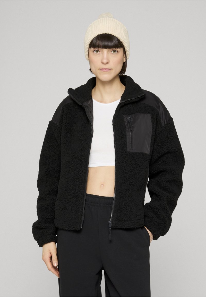 Černá fleecová bunda se zapínáním na zip vpředu a texturovaným povrchem. Má černé nylonové panely a zipovou kapsu na hrudi. Měkký, ležérní střih.