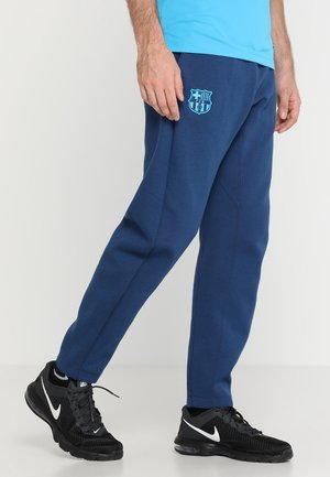 Homme portant un pantalon de survêtement bleu du FC Barcelone et des baskets noires Nike, debout sur un sol blanc devant un fond uni.