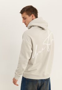 Mladi moški, obrnjen v stran, nosi svetlo bež jopa s kapuco, na hrbtni strani je napisano "Your Sun", in modre jeans hlače.