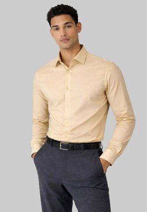 Homme portant une chemise beige à manches longues boutonnée rentrée dans un pantalon gris foncé avec une ceinture noire, debout les mains dans les poches.