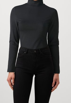 Femme portant un haut noir moulant à col roulé et manches longues, et un jean noir taille haute, debout devant un fond clair uni.