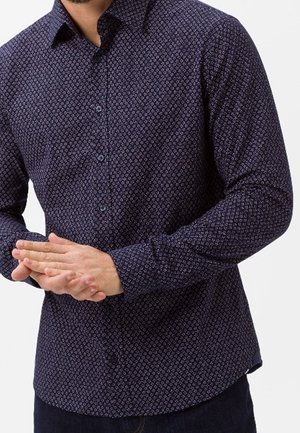 Mann trägt ein dunkelblaues langärmeliges Hemd mit kleinen weißen geometrischen Mustern, die Hände vor dem Körper verschränkt.