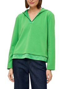 Zielona bluza z kapturem w kształcie V; długie rękawy i gładka tekstura. Posiada warstwowy dół i luźny fason. Połączona z ciemnymi dżinsami.