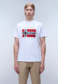 Bílý bavlněný tričko s barevným grafickým designem vlajek a textem "Napapijri Geographic". Model v béžových kalhotách.