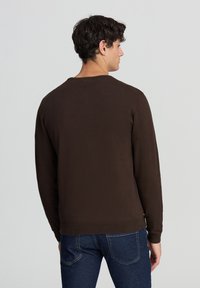 Maglione marrone lavorato a maglia con scollatura rotonda, maniche lunghe e polsini e orlo a coste. Indossato sopra jeans blu, mostrato da dietro.