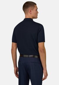 Homme de profil portant un polo bleu marine à manches courtes, un pantalon de ville bleu foncé et une ceinture noire, sur un fond uni.