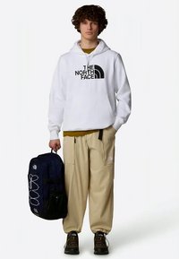 Bela kapuca s kapuco z črnim logotipom, bež cargo hlače z žepi, črn in mornarsko moder nahrbtnik ter temni čevlji. Sproščena oblačila za zunaj.