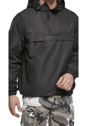 Homme portant un coupe-vent noir à capuche avec des poches zippées et un pantalon cargo camouflage gris, debout avec une main légèrement levée.