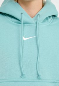 Un hoodie bleu clair en tissu doux, arborant un logo Nike blanc sur la poitrine et deux cordons assortis avec des pompons aux extrémités.