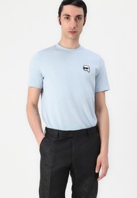 Light blue short-sleeved t-shirt with a round neck and a small black and white graphic patch on the chest, paired with black patterned trousers.
