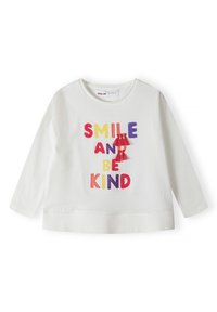 Weißes Langarmshirt aus Baumwolle mit buntem, gesticktem Text, der "LÄCHEL UND SEI NETT" liest, mit roten Quastenakzenten.