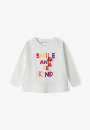 Vit långärmad skjorta av bomull med färgglad, broderad text som säger "LE ENDAST OCH VAR SNÄLL," med röda tofsar som accenter.