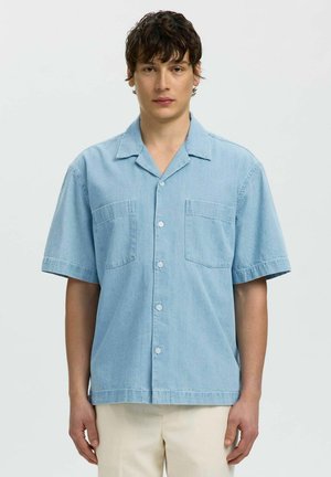 Jeune homme portant une chemise bleu clair à manches courtes avec deux poches poitrine, debout devant un fond blanc uni.
