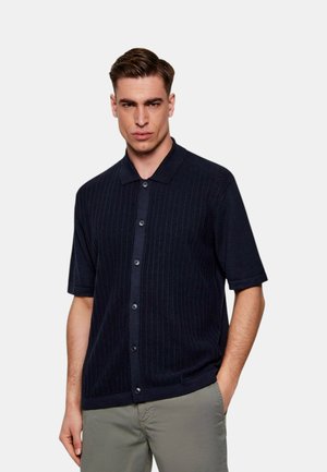 Uomo che indossa una camicia blu navy a maniche corte abbottonata con sottili righe verticali e pantaloni grigio chiaro su uno sfondo bianco.