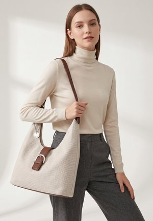 Vrouw in een beige coltrui en grijze broek houdt een grote beige handtas met een bruine band en gesp over haar schouder tegen een effen achtergrond.