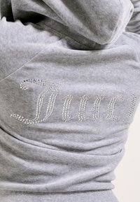 Siva dukserica od mekanog materijala s sjajnim kamenčićima koji oblikuju riječ "Juicy" na leđima.