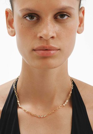 Primer plano de una mujer que lleva un collar de cadena dorada con piedras preciosas redondas y transparentes, vestida con un top halter negro sobre un fondo blanco.