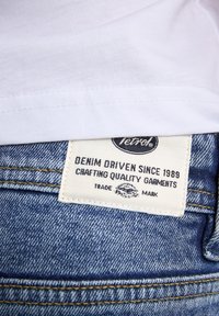 Biała bawełniana koszulka z szwem na dole, nałożona na niebieskie dżinsy. Dżinsy mają białą metkę z czarnym tekstem i żółtymi przeszyciami.