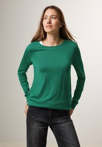 Grünes Langarmshirt mit rundem Ausschnitt, aus glattem Stoff, kombiniert mit dunkelgrauen Jeans. Einfaches und körpernahes Design.