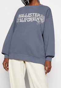 Blå oversized sweatshirt i bomullsblandning, med tryck "HOLLISTER CALIFORNIA" i vitt, raglanärmar och ribbade ärmslut.