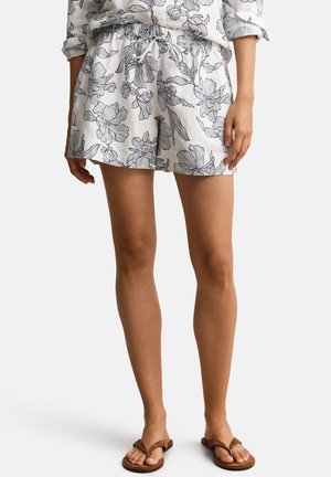 Frau trägt weiße Shorts mit schwarzem Blumenmuster und braunen Sandalen, steht vor einem schlichten hellen Hintergrund.