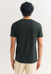 Temno zelena majica s kratkimi rokavi iz gladkega blaga, z okroglim izrezom in ravnim krojem, nošena s svetlo obarvanimi hlačami.