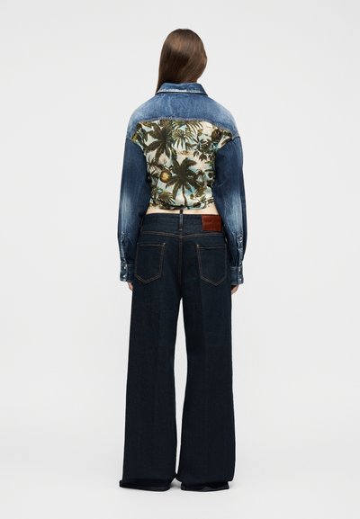 Γυναίκα που φοράει τζιν μπουφάν με τροπικό μοτίβο φοινικόδεντρου στην πλάτη και σκούρο τζιν με φαρδιές φάσες, στέκεται γυρισμένη πλάτη σε λευκό φόντο.