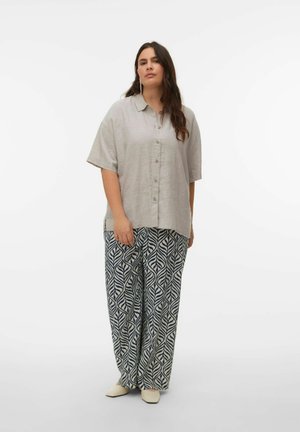 Frau trägt locker sitzendes beiges Hemd mit Knöpfen, gemusterte weite Hosen und cremefarbene Schuhe, steht vor einem schlichten weißen Hintergrund.