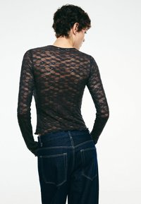 Haut à manches longues en dentelle noire avec un motif de fleurs texturé, ourlet festonné, associé à un jean bleu foncé. La vue montre l'arrière de la tenue.