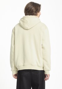 Sudadera color crema con un interior texturizado y forrado de felpa. Presenta una capucha con cordón, puños acanalados y dobladillo. Combinada con pantalones negros.