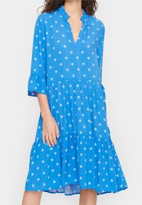 Blå prickig klänning i lättviktig tyg, har V-ringning, volangärmar och en flerskiktad kjol med en flytande silhuett.